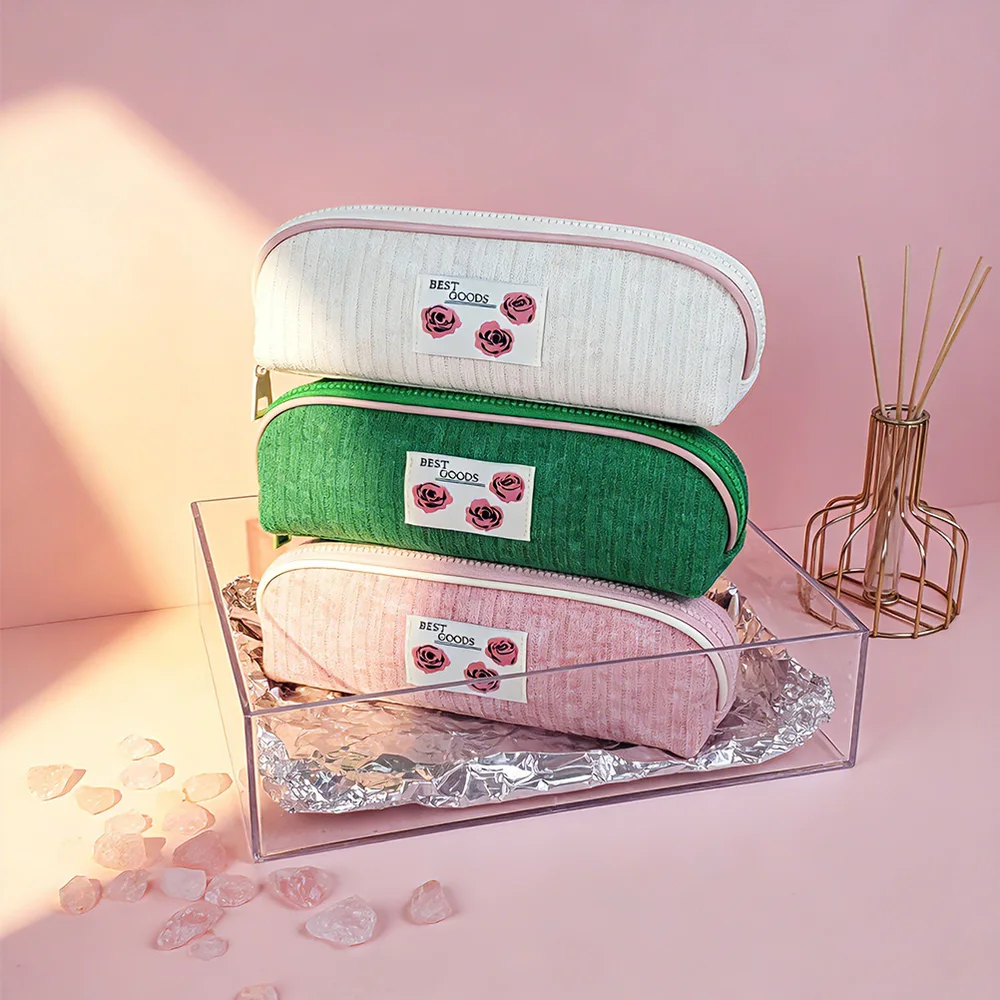 Three corduroy barrel pouches in cream, green, and pink stacked on a clear acrylic box against a pink backdrop with rose quartz crystals and a gold reed-diffuser vase