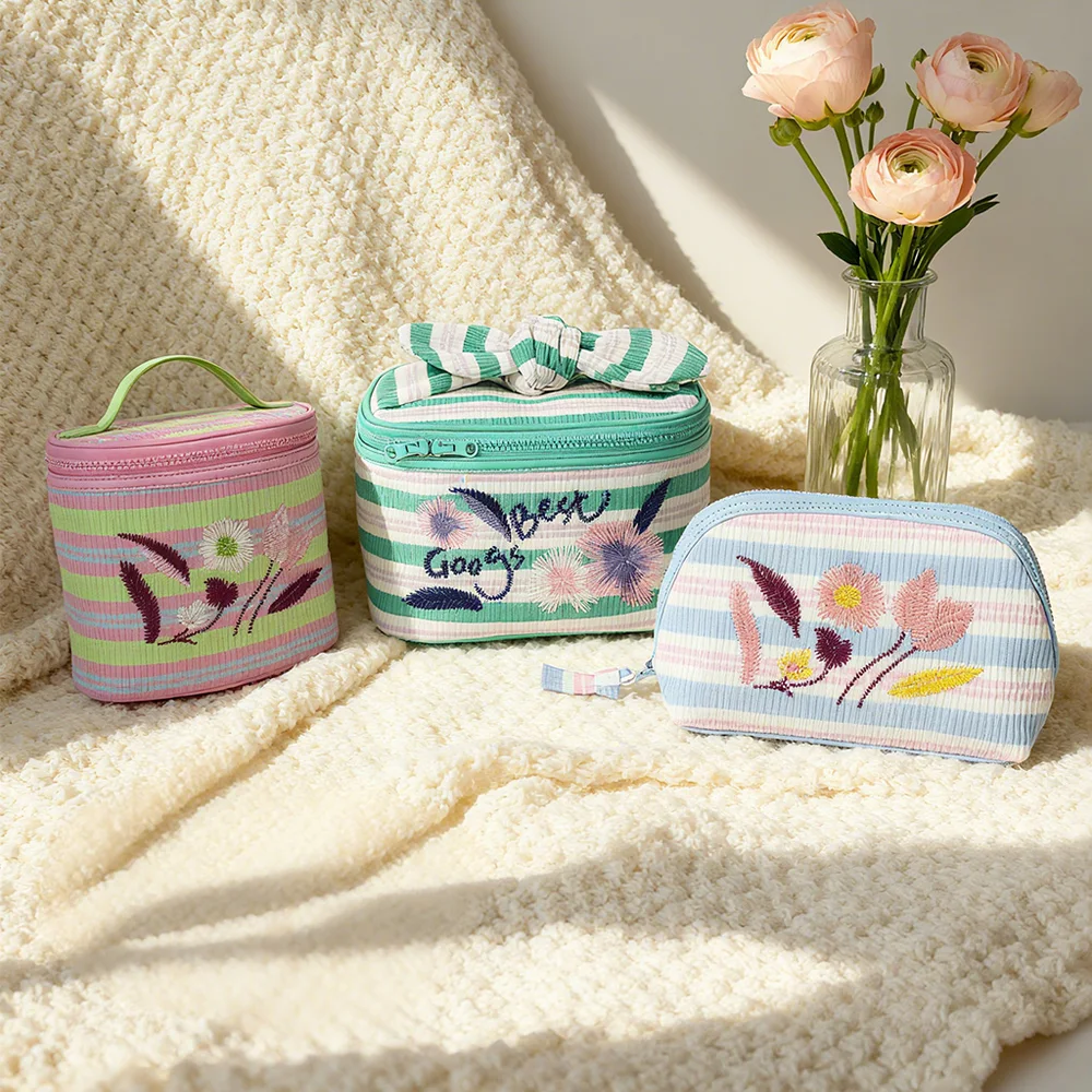 Three pastel striped corduroy cosmetic pouches with embroidered flowers styled on a cream knit blanket beside a glass vase of peach ranunculus