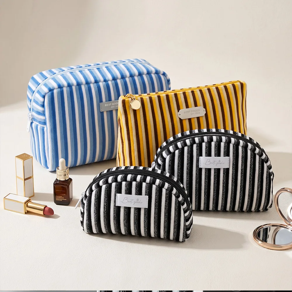 Four striped corduroy cosmetic bags in blue, mustard, and black-and-white arranged on a cream surface with lipstick, serum, and compact mirror props