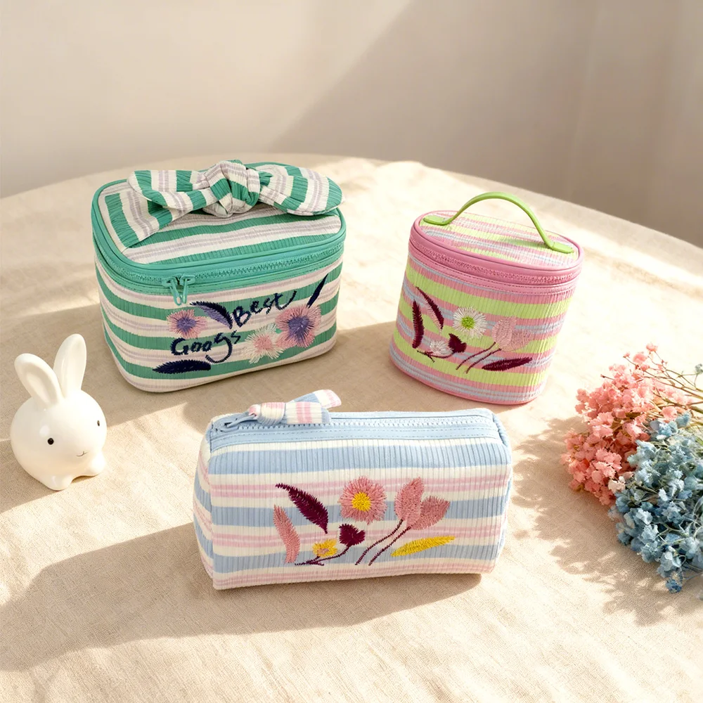 Three pastel striped corduroy cosmetic bags with floral embroidery, knotted bow detail and green top handle, styled on beige cloth with ceramic bunny and dried flowers