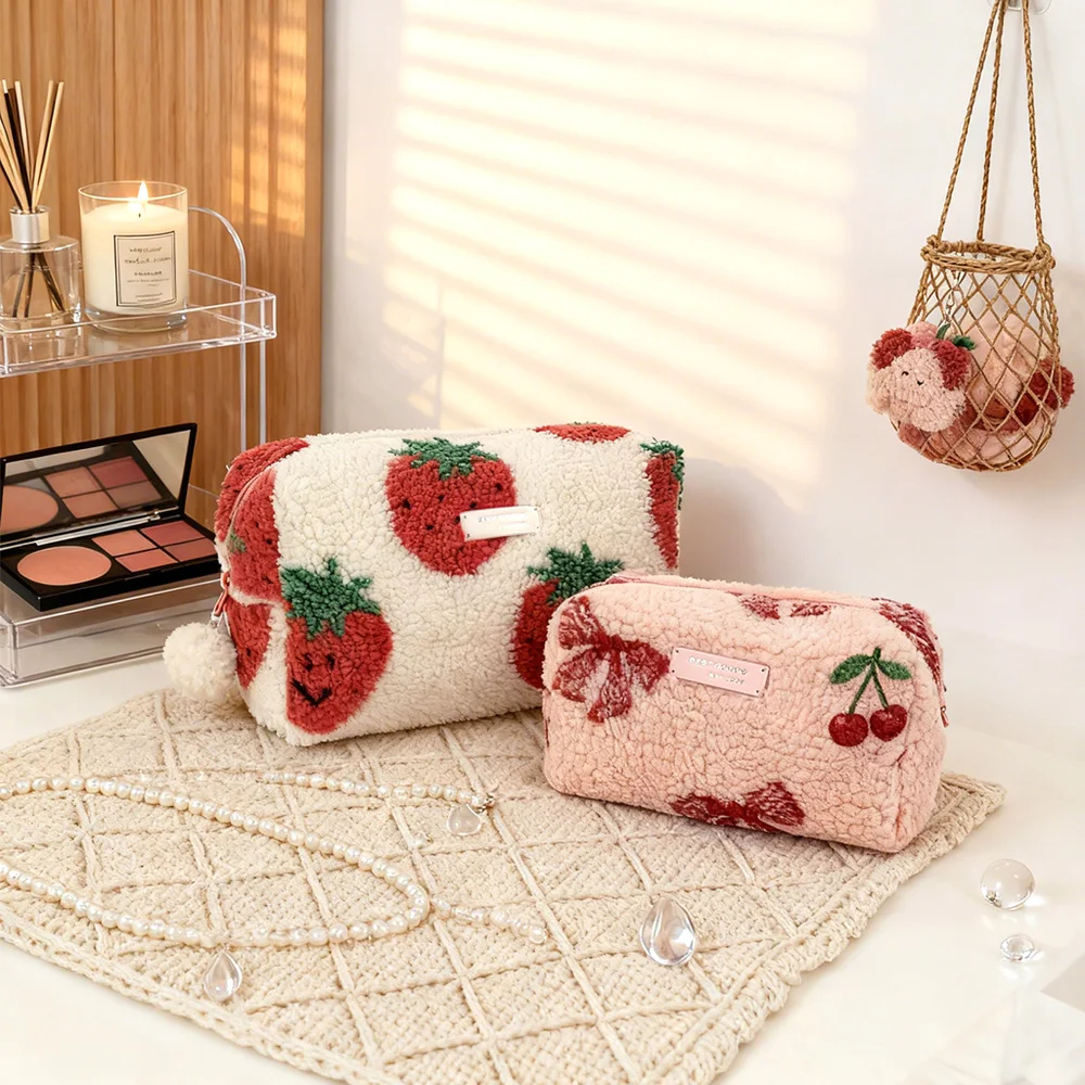Two sherpa fleece cosmetic pouches, one cream with tufted smiling strawberries, one blush pink with red bows and cherry, styled on woven mat with candle, makeup palette and rattan basket