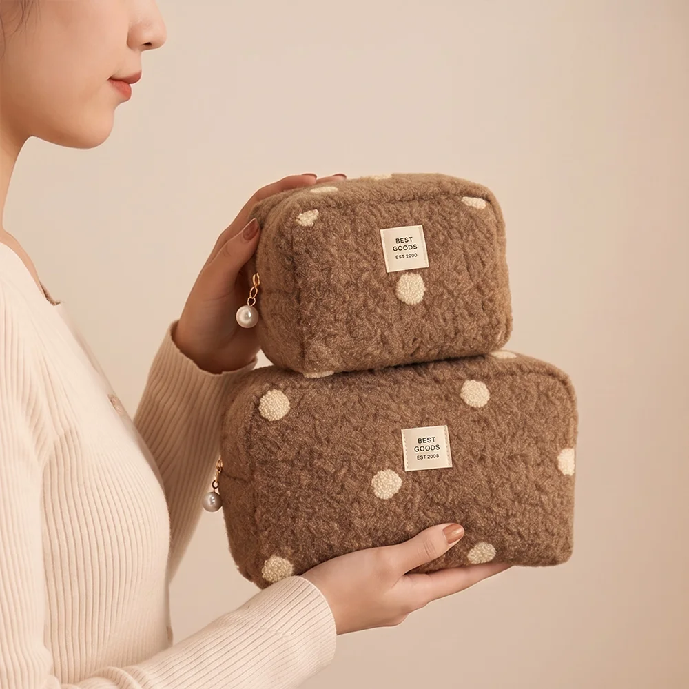 Two camel-brown sherpa polka-dot boxy cosmetic pouches with gold zipper pulls and pearl charms, stacked and held by a model in a cream ribbed sweater against beige backdrop