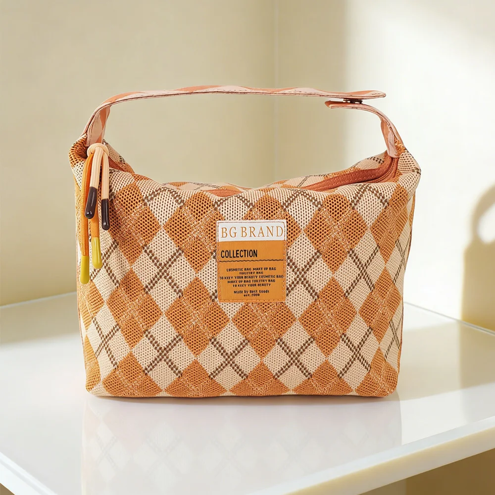Orange and cream argyle-knit cosmetic pouch with wide top zipper, peach handle, and BG BRAND woven label, on white table against cream wall