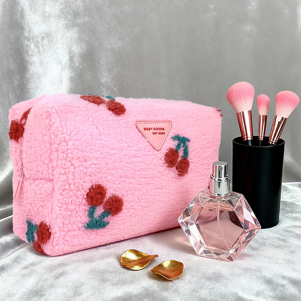 Pink sherpa fleece zippered makeup pouch with embroidered cherries and BEST GOODS triangle patch, styled with diamond-shaped perfume bottle and pink makeup brushes in black holder on silver satin backdrop
