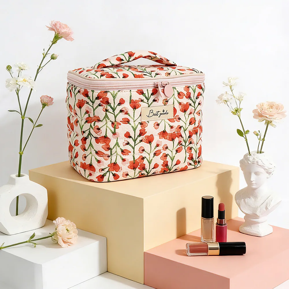 Quilted cream cosmetic bag with red poppy floral print and blush-pink zipper, styled on pastel pedestals with flowers, a bust vase, and lip products