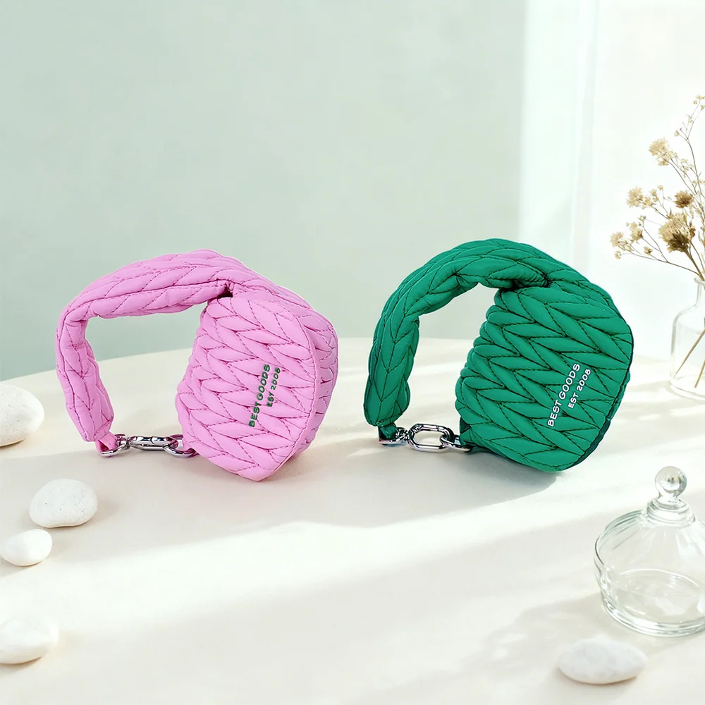 Pink and emerald green quilted puffer mini pouches with braided loop handles and silver chain links on white fabric with pebbles, glass jar, and dried flowers