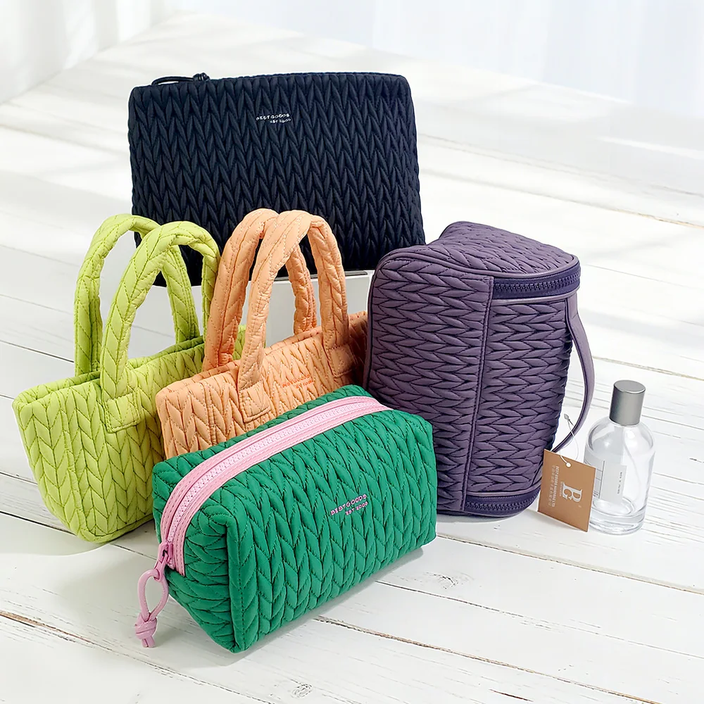 Set of five chevron-quilted puffer cosmetic bags in navy, lime, peach, green, and purple arranged on white wood floor with perfume bottle and brand tag