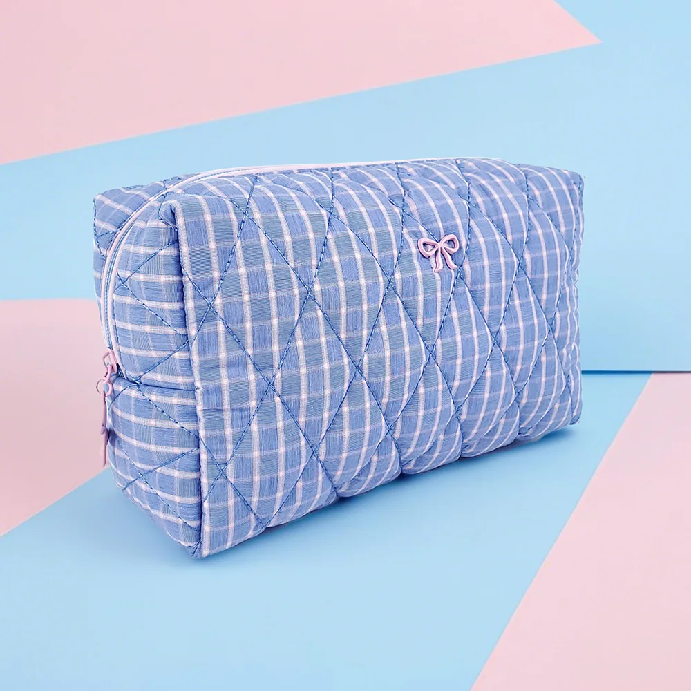 Blue gingham diamond-quilted puffy cosmetic pouch with pink bow detail and pink cord zipper pull, set against pink and light-blue geometric backdrop