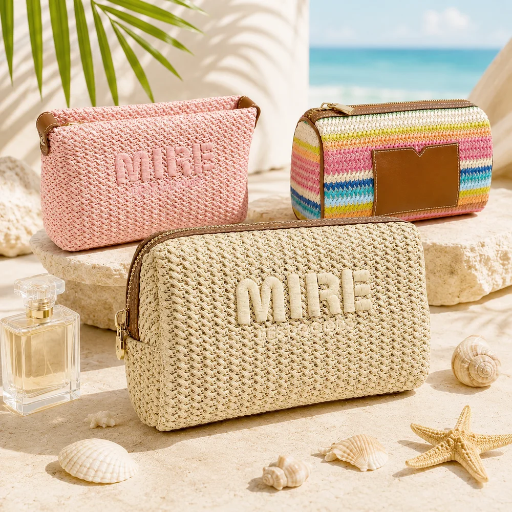 Three woven straw MIRE cosmetic pouches in natural, blush pink, and rainbow stripe styled on stone pedestals with seashells, starfish, perfume bottle, and ocean backdrop