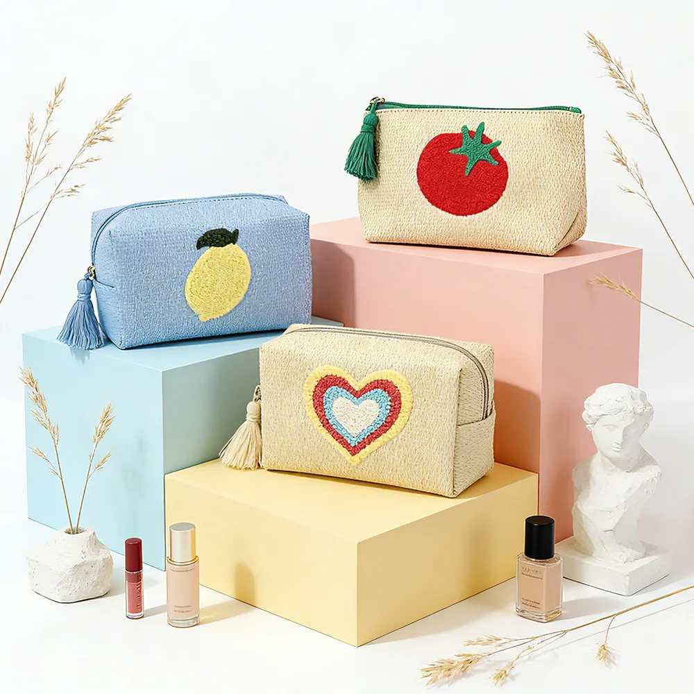Three straw-texture zip pouches with embroidered lemon, tomato, and rainbow-heart motifs staged on pastel pink, blue, and yellow blocks with dried grasses, a small bust, and cosmetic bottles