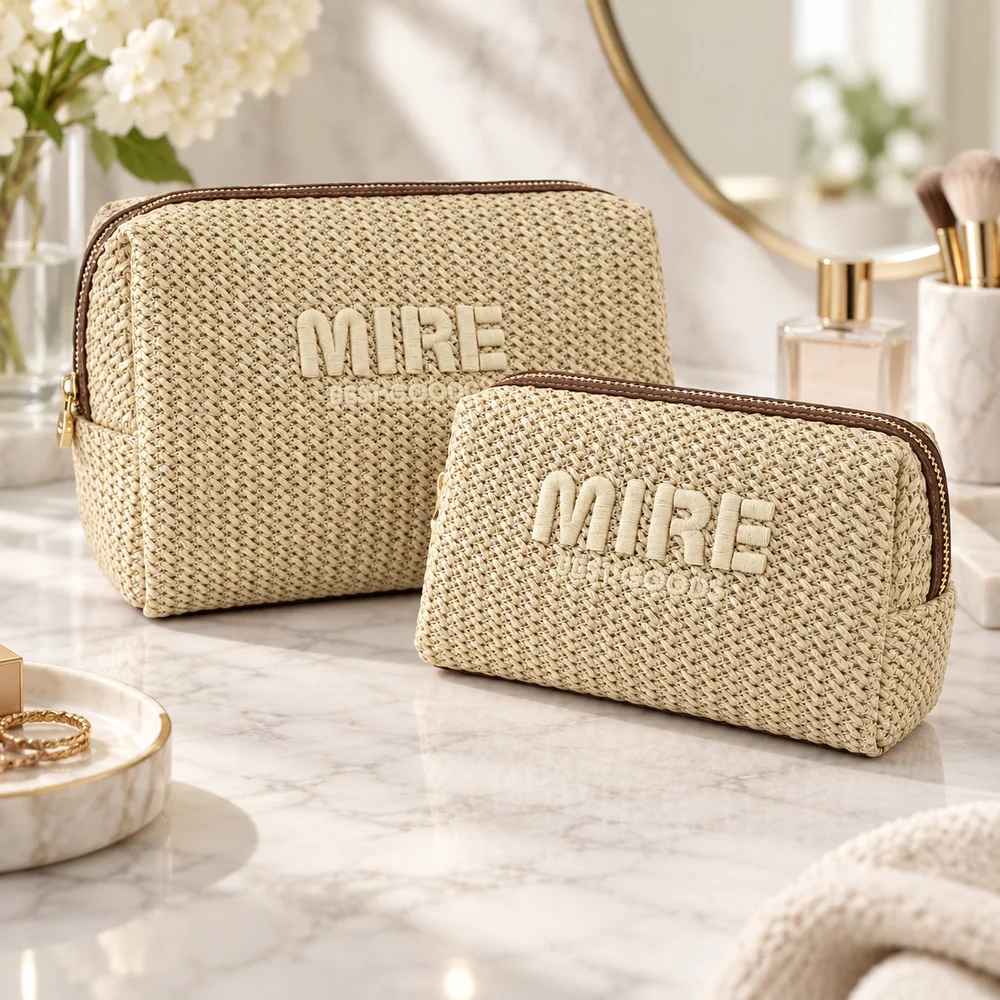 Two natural chevron-straw zip pouches with brown zippers, gold pulls, and raised MIRE embroidery on a marble vanity with gold mirror, perfume, brushes, and flowers
