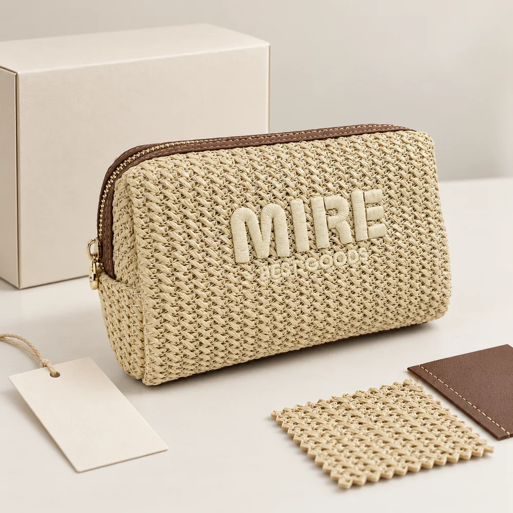 Natural straw-woven cosmetic pouch with brown leather-trim gold zipper and raised MIRE embroidery, styled with cream gift box, paper hang tag, and straw and leather swatches on beige surface