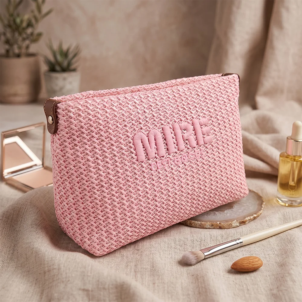 Blush-pink woven raffia cosmetic pouch with raised MIRE embroidery styled on beige linen with mirror, serum bottle, makeup brush and almond props