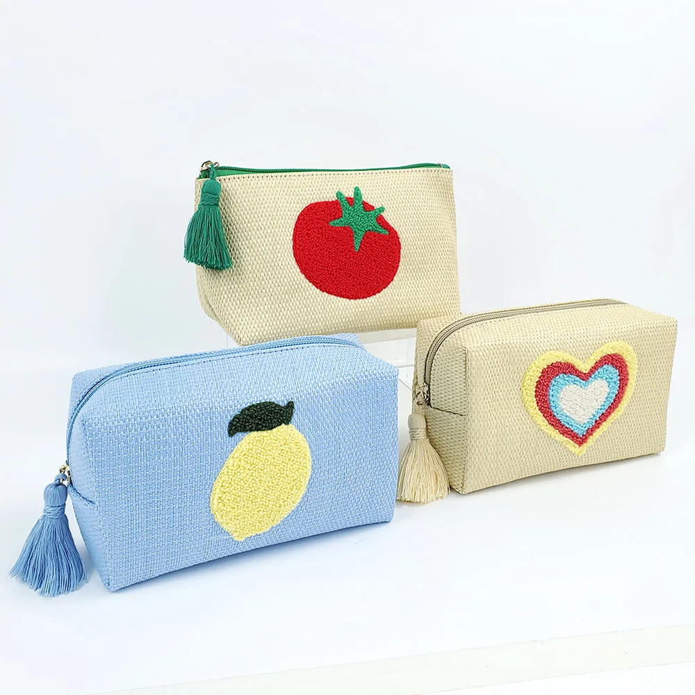 Three woven straw zipper pouches with embroidered tomato, lemon, and rainbow-heart motifs and tassel pulls, arranged on white background