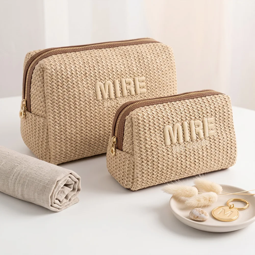 Two champagne woven straw zippered pouches with MIRE logo and gold hardware on white table, rolled linen towel and ceramic dish with dried flowers and gold charm nearby