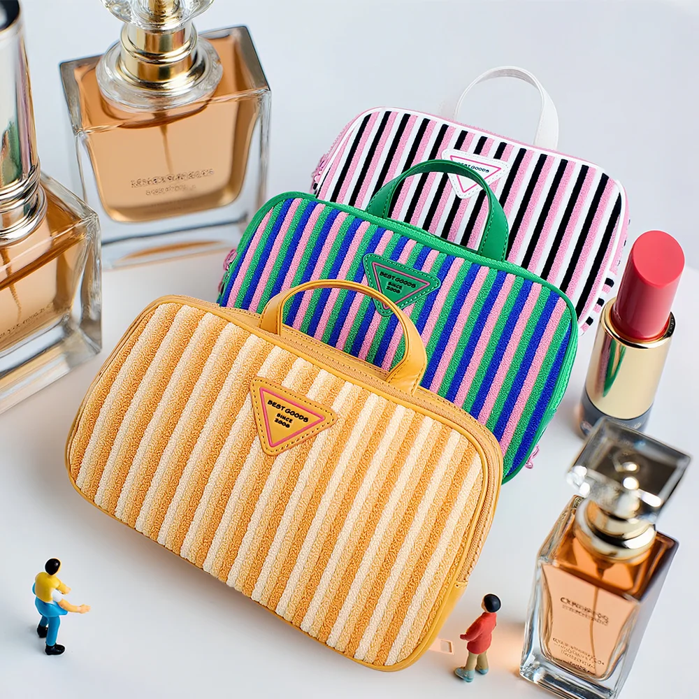 Three striped terry cloth cosmetic pouches in butter-yellow, green-blue-pink, and pink-white-black with top handles and triangular BEST GOODS patches, styled with perfume bottles, a gold lipstick, and miniature figures on a white surface
