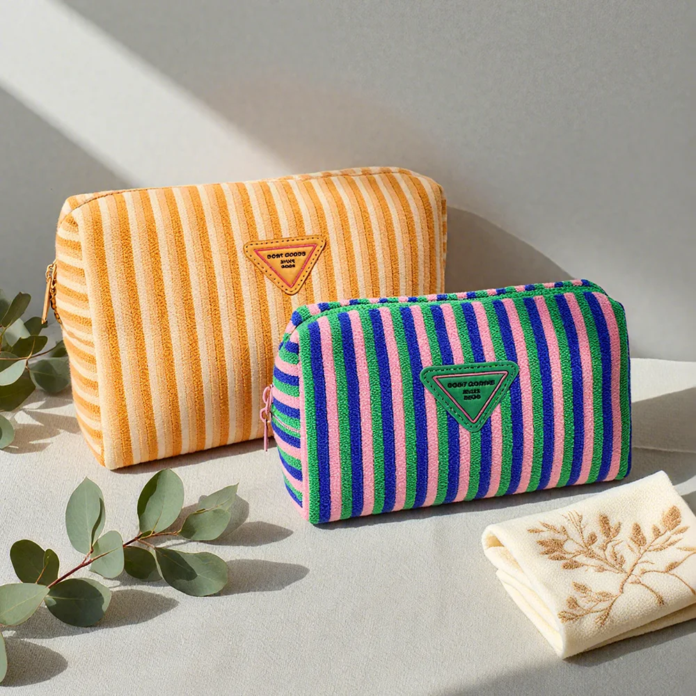 Two striped terry cloth zip pouches, one orange and cream, one pink, blue and green, with triangle logo patches on a linen surface beside eucalyptus and an embroidered cloth