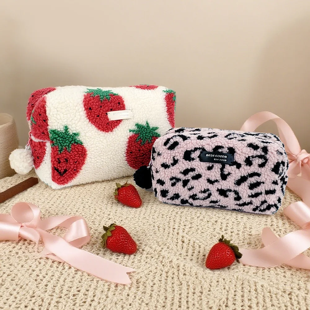 Two plush tufted cosmetic bags, cream with red strawberries and blush-pink with black leopard print, styled on a knit blanket with fresh strawberries and pink satin ribbons