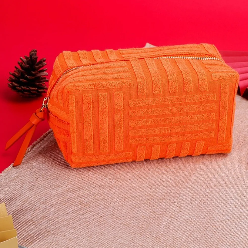 Orange terry-towel box cosmetic pouch with gold zipper and leather pull tab resting on a cream woven mat against a bright red background with pinecone prop