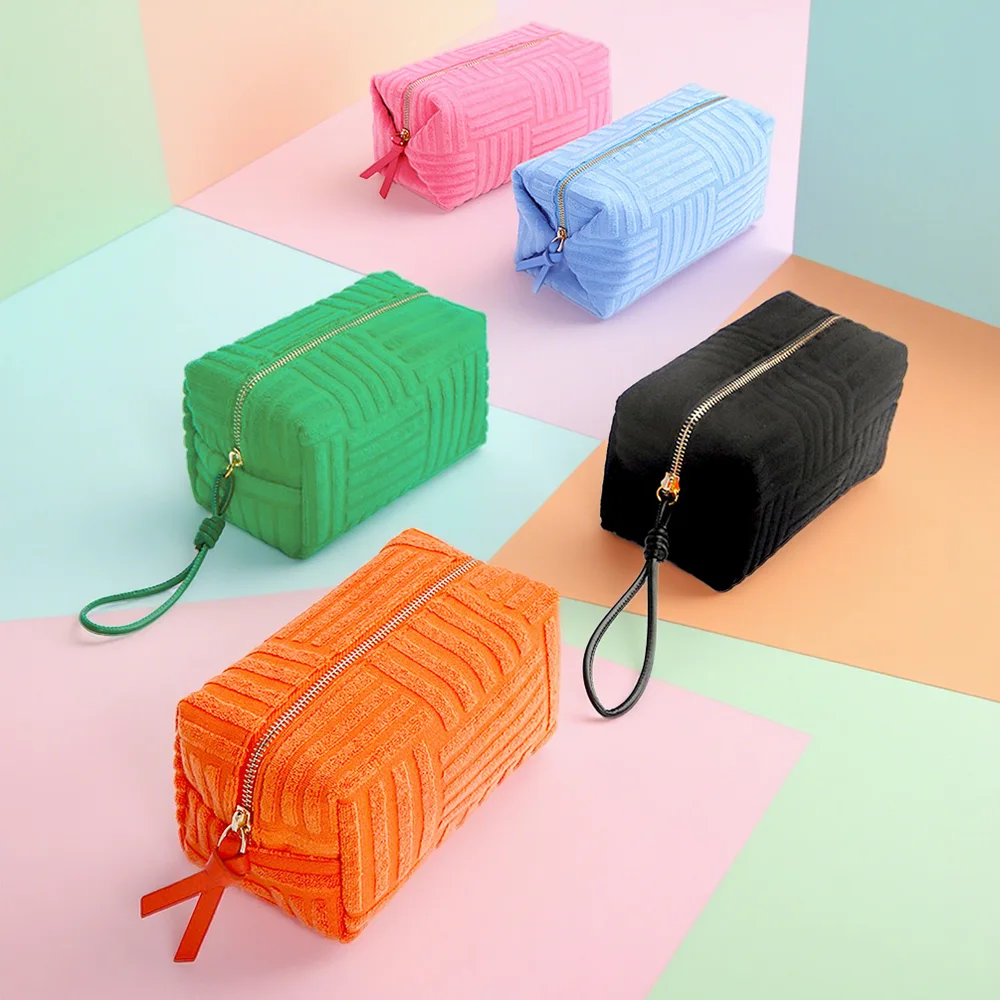 Five quilted terrycloth box pouches in pink, blue, green, black, and orange with ribbon zip pulls on pastel paper backdrop