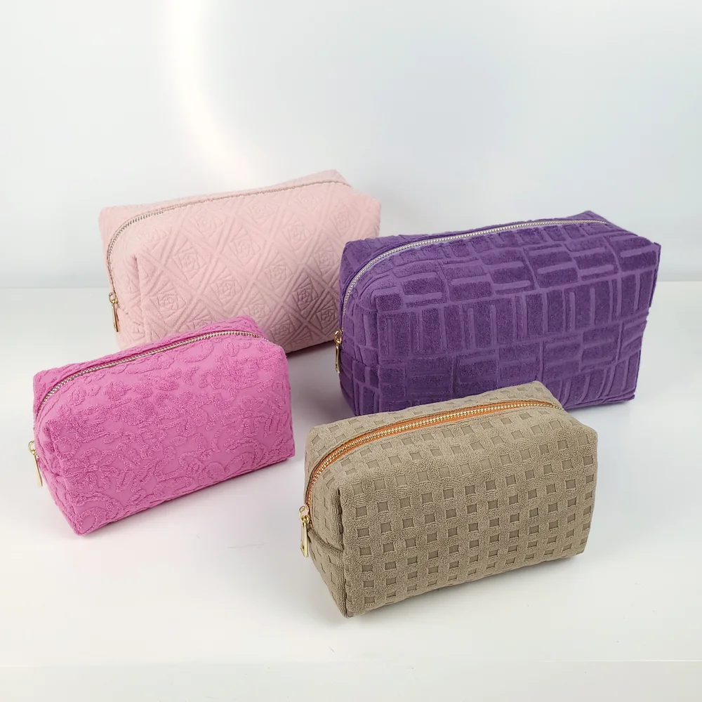 Four terry towel box cosmetic bags in blush pink, magenta, purple and taupe with embossed textures and gold zippers on white background