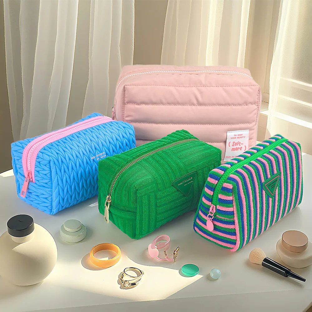 Four pastel puffer, braided, and terry cloth cosmetic pouches in pink, blue, and green styled on a sunlit white table with jewelry and makeup props