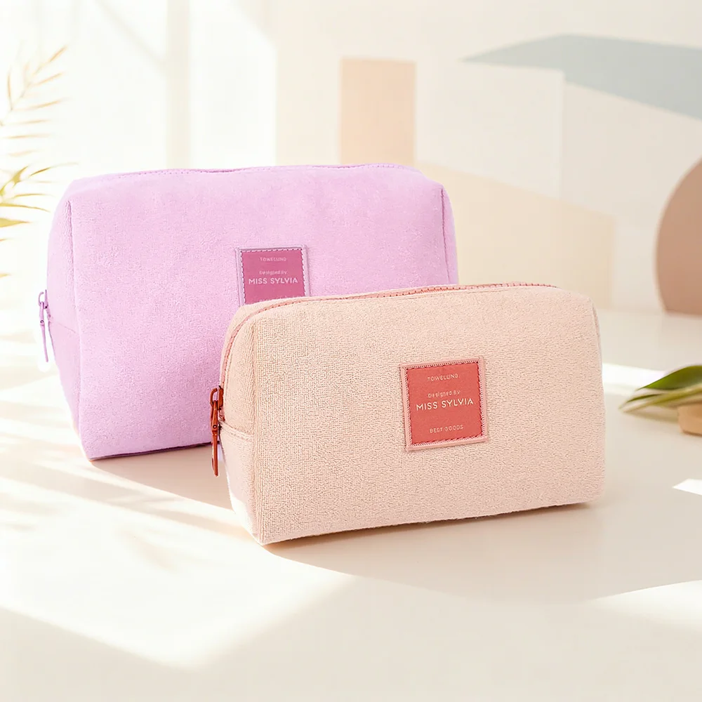 Two terry cloth zippered cosmetic pouches in blush pink and lavender with red MISS SYLVIA woven labels, styled on a pastel tabletop with plant leaf