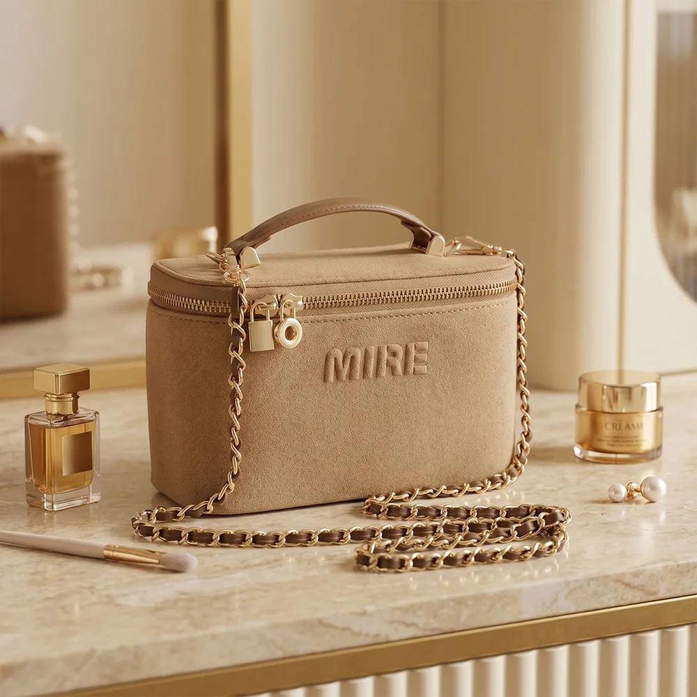 Tan camel suede vanity case with gold chain strap, top handle, and debossed MIRE logo on marble vanity with perfume bottle, cream jar, and pearl earrings