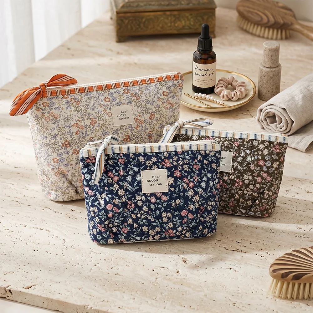 Three quilted floral zip pouches in navy, olive, and cream with ribbon bow zipper pulls arranged on travertine stone beside skincare oil, brushes, and towel