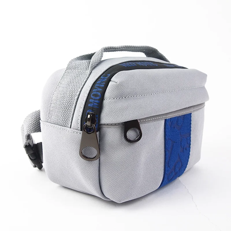 Light gray box-shaped cosmetic pouch with royal blue woven front stripe, wide top handle, shoulder strap, and black metal zipper pulls, on white background