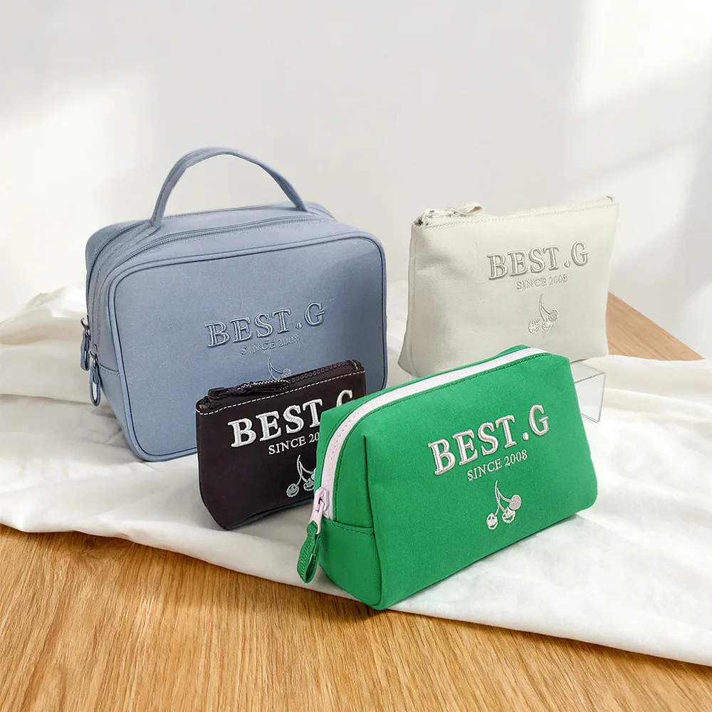 Four canvas zip cosmetic bags in dusty blue, cream, black and green with BEST.G SINCE 2008 cherry embroidery arranged on white cloth over wooden table