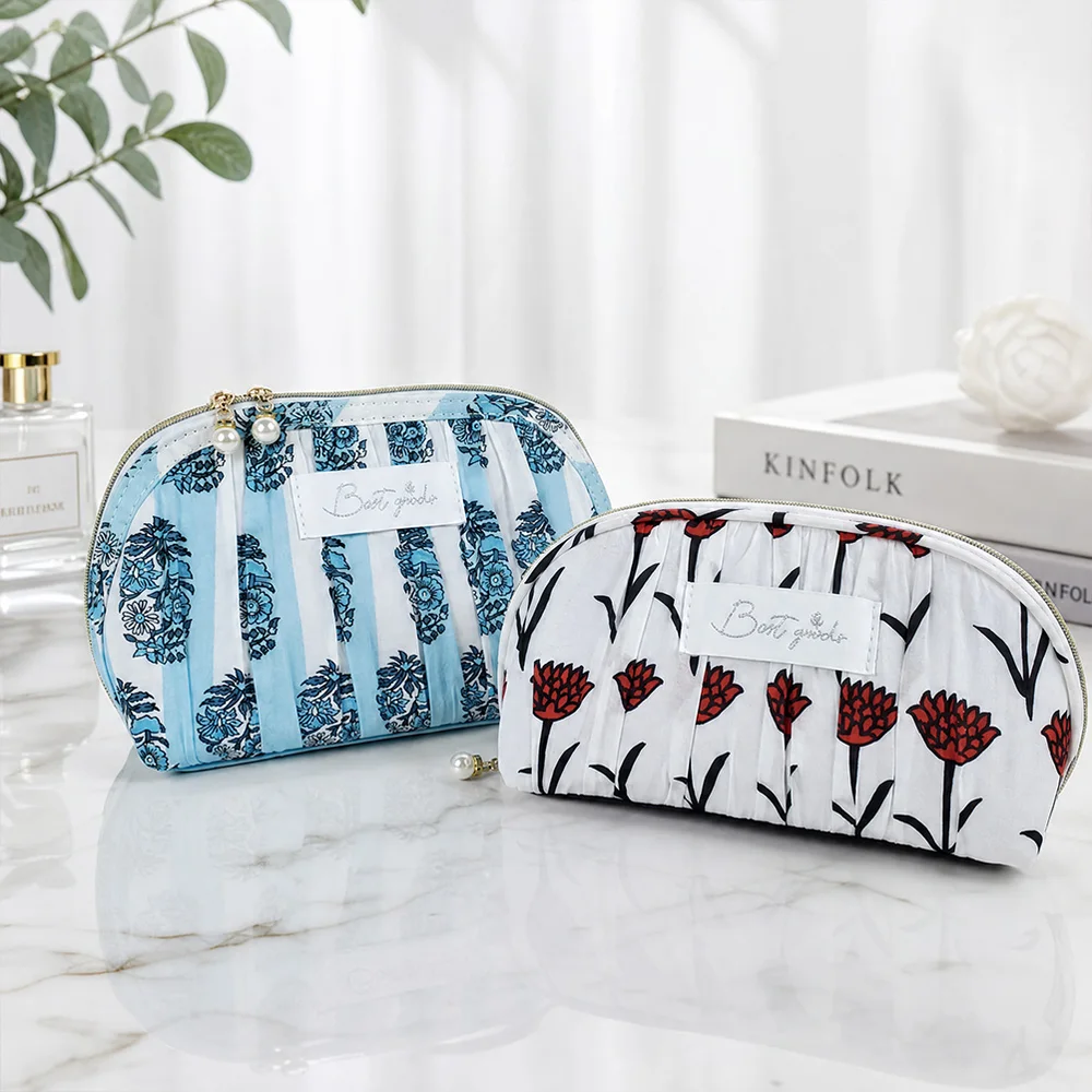 Two pleated dome cosmetic pouches with pearl zipper pulls, one blue floral and one red poppy print, on white marble beside perfume, books, and flowers