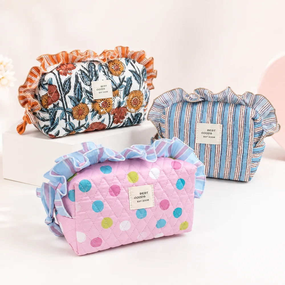 Three quilted cotton cosmetic pouches with ruffle-trim tops and woven BEST GOODS labels: orange floral, blue-pink stripe, and pink polka dot, arranged on white pedestal against cream backdrop