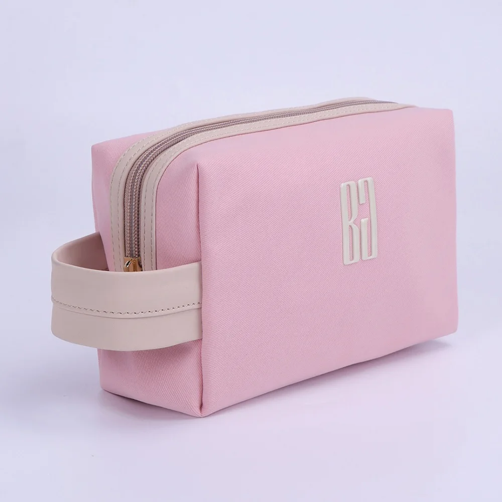Blush-pink rectangular cosmetic bag with cream top handle, gold zipper, and BG metal logo plaque on light background