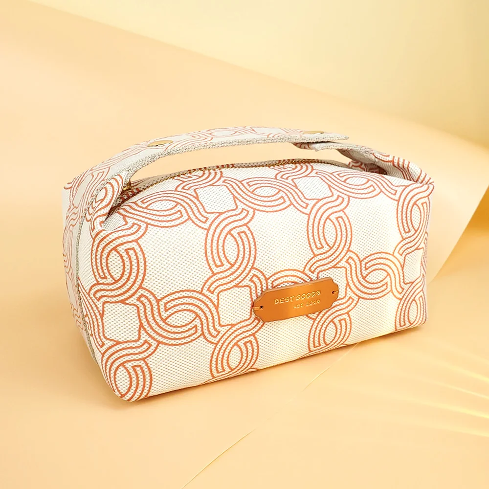 Ivory canvas boxy cosmetic bag with rust-orange geometric chain print, gold-riveted top handle and copper BEST GOODS metal plate, on peach paper backdrop