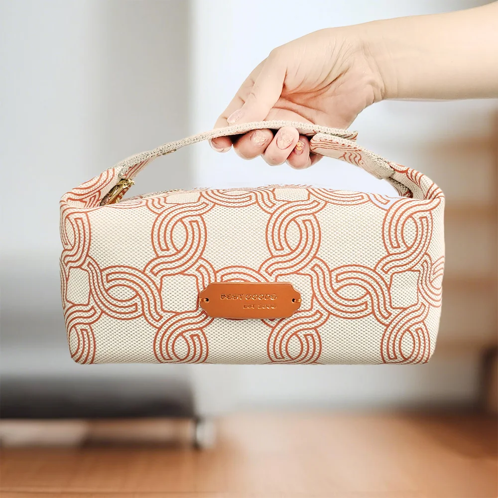 Hand holding a cream canvas makeup pouch with terracotta geometric monogram print, tan leather logo patch, and gold zipper against a blurred interior with wood floor