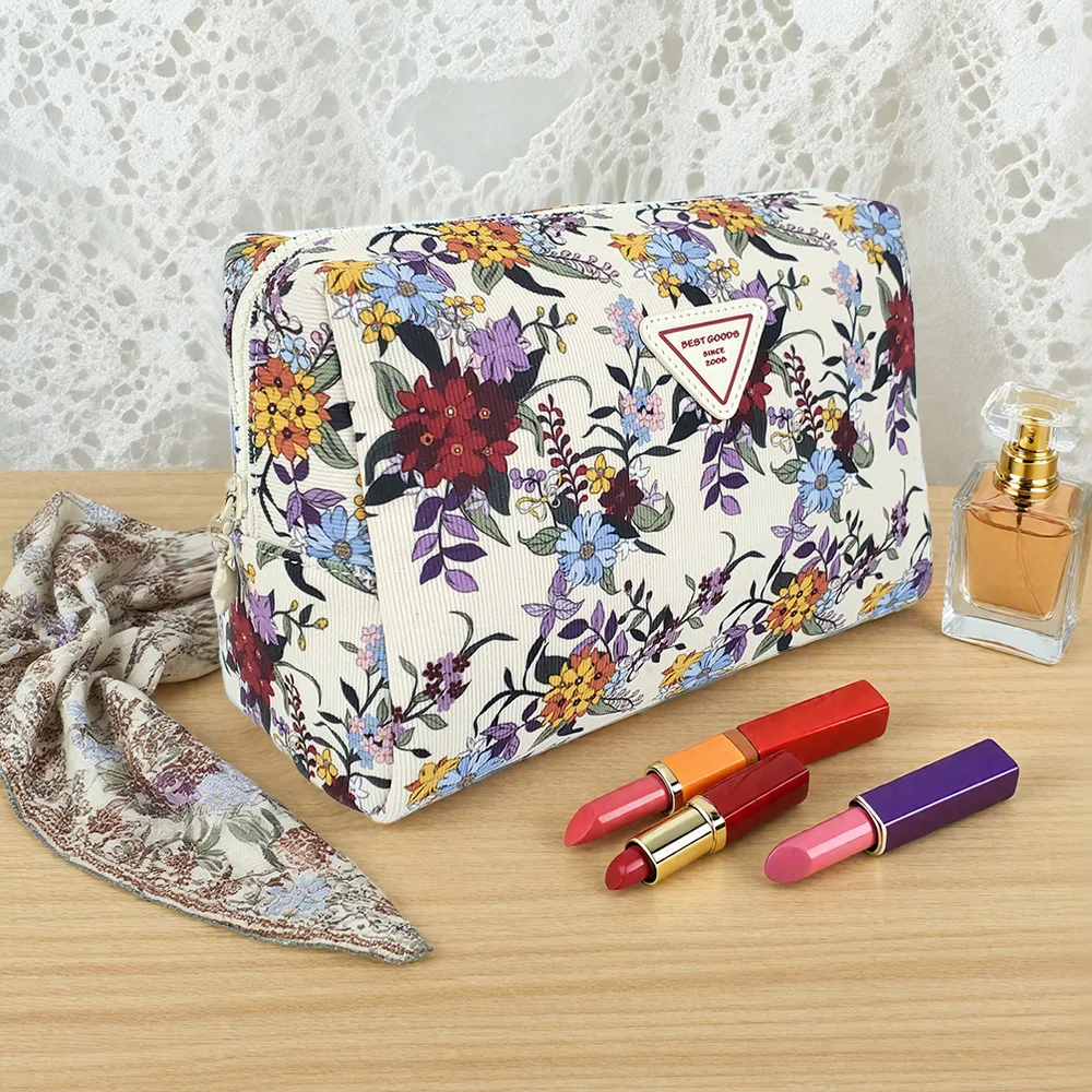 Cream floral-print trapezoid cosmetic pouch with white triangle logo patch on light wood table, styled with perfume bottle, three lipsticks, and paisley scarf against white lace backdrop