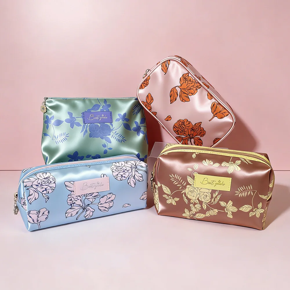 Four satin floral-print cosmetic pouches in sage green, blush pink, powder blue, and copper arranged on a pink seamless backdrop