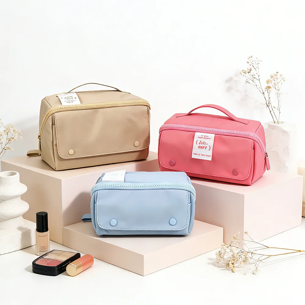 Three rectangular zip-top cosmetic bags with top handles in tan, coral pink, and powder blue displayed on pink pedestals with dried flowers and makeup props