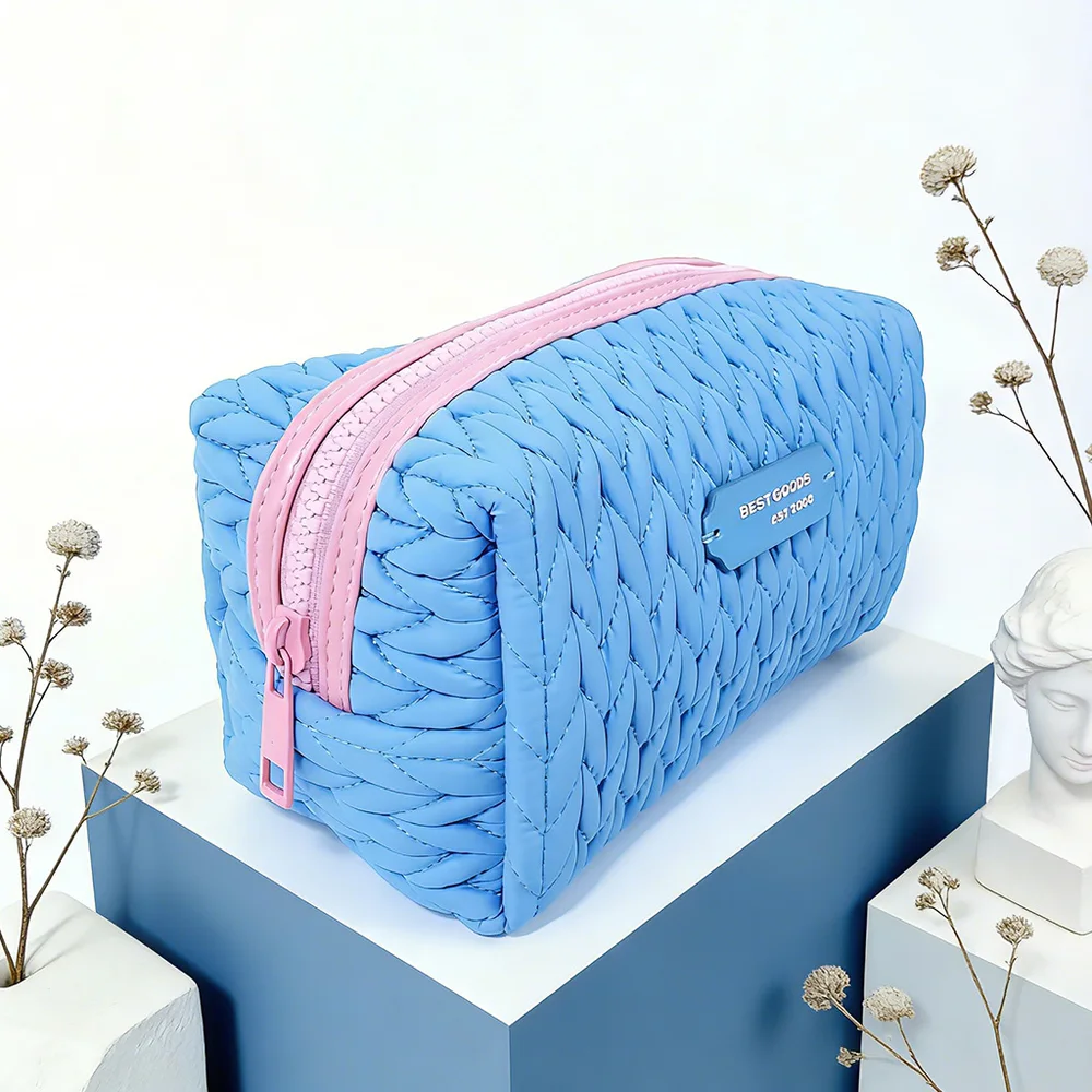 Cornflower-blue braided quilted box makeup pouch with pink zipper, styled on white and blue display blocks with dried flowers and white bust statue