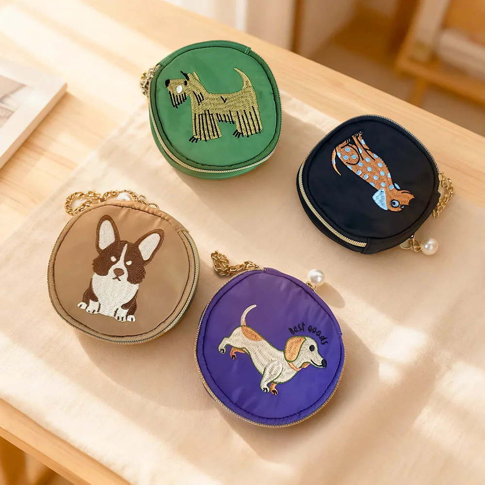 Four round nylon coin pouches in green, navy, tan, and purple with embroidered dog motifs, gold perimeter zippers, chain wrist straps, and pearl charms on a wooden table with cream cloth