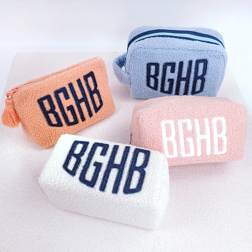 Four sherpa cosmetic pouches in coral, powder blue, blush pink, and ivory with embroidered BGHB monogram arranged on pale pink surface