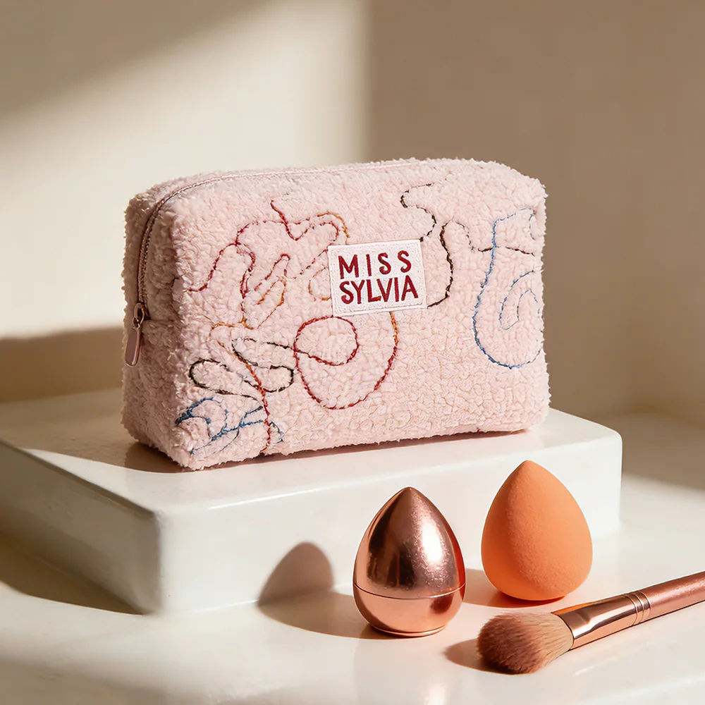 Blush-pink sherpa cosmetic pouch with MISS SYLVIA woven label and abstract embroidery on white pedestal with rose-gold brush and makeup sponges