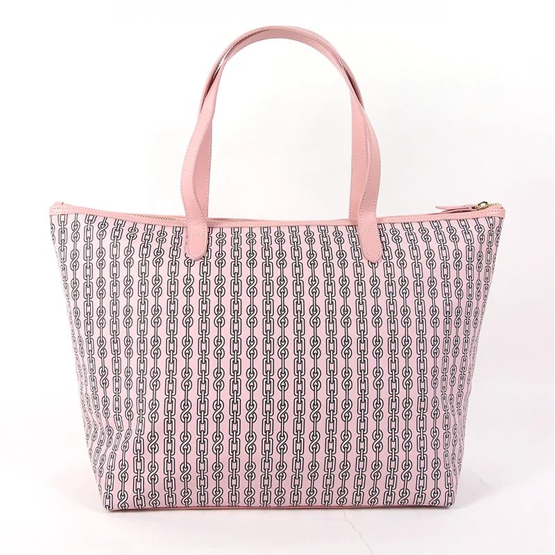 Blush pink zip-top tote with navy chain-link print, tonal monogram center stripe, pink leather-look top handles, and gold zipper pull on white studio background