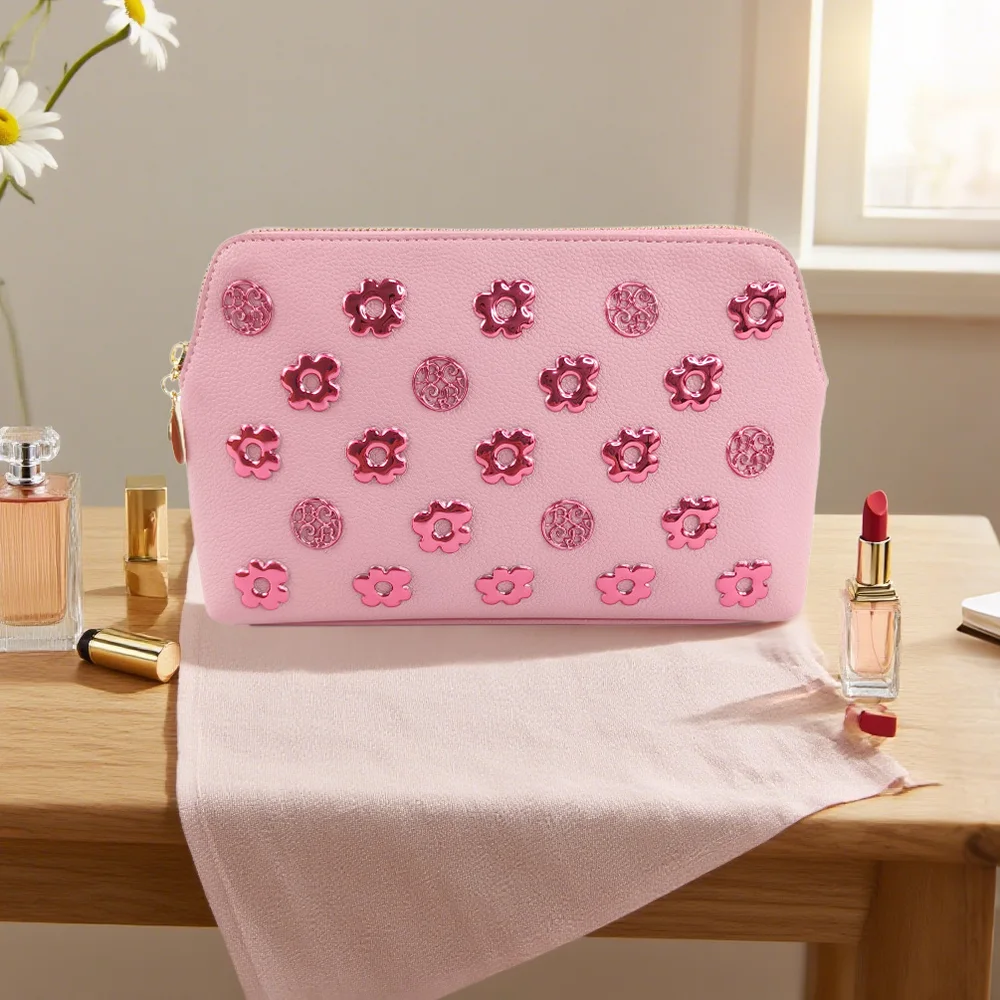 Light pink PU zipper pouch decorated with pink metallic flower and embossed disc appliqués, on wooden table with perfume bottles, red lipstick, and white daisies by a window