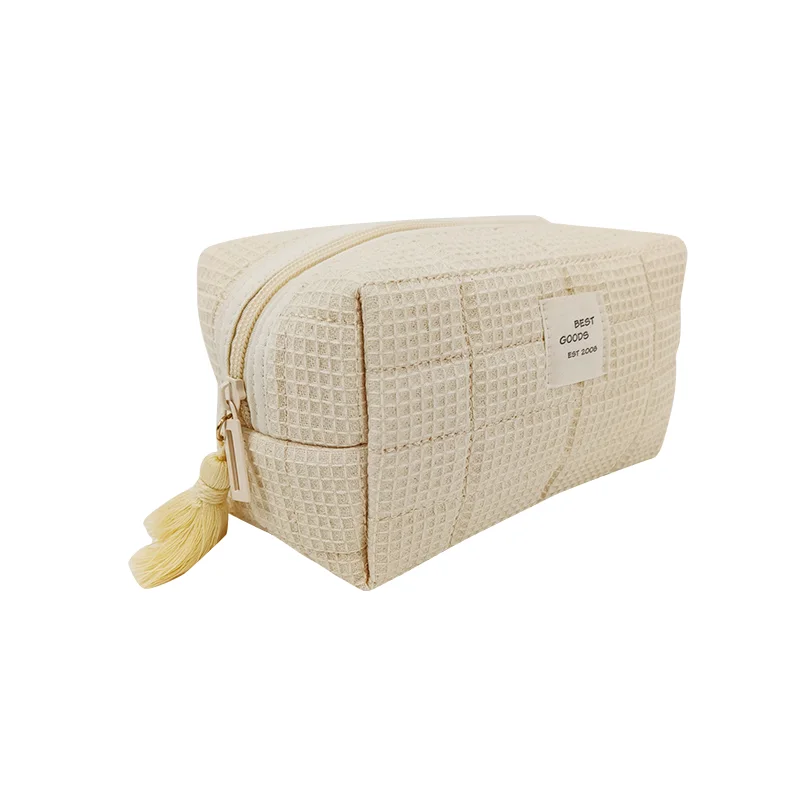 Cream waffle-weave boxy cosmetic pouch with top zipper, cream pull, and butter-yellow tassel on a white background
