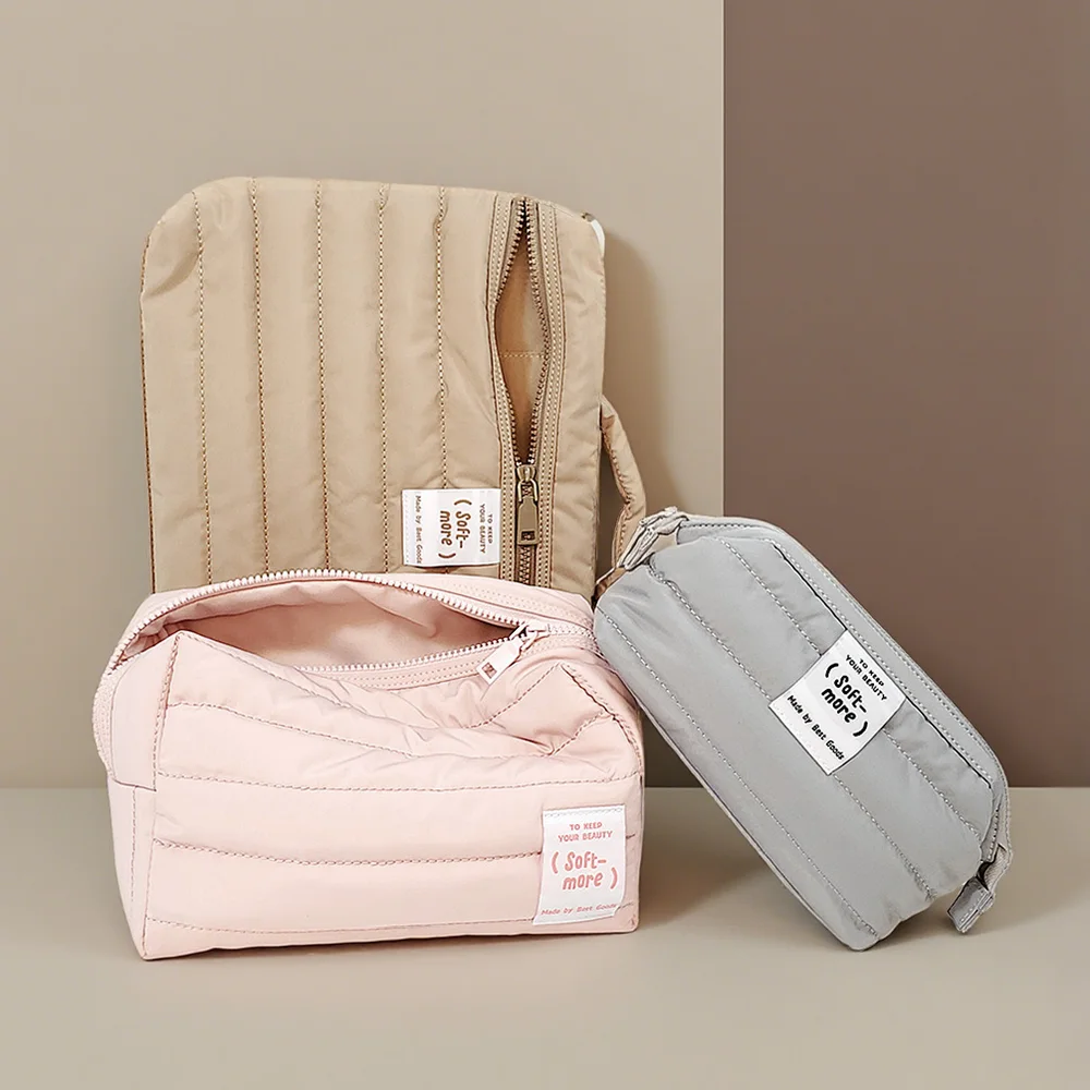 Three quilted puffer cosmetic bags in beige, blush pink and grey with woven Soft-more labels on a two-tone taupe studio background