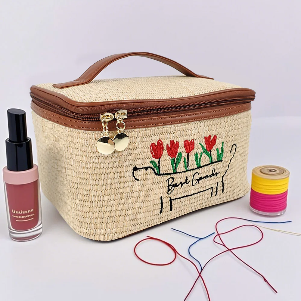 Natural woven straw box-shaped cosmetic case with cognac top handle, gold dual zipper pulls, and red tulip embroidery reading Best Goods, styled with nail polish and thread spools on a white background