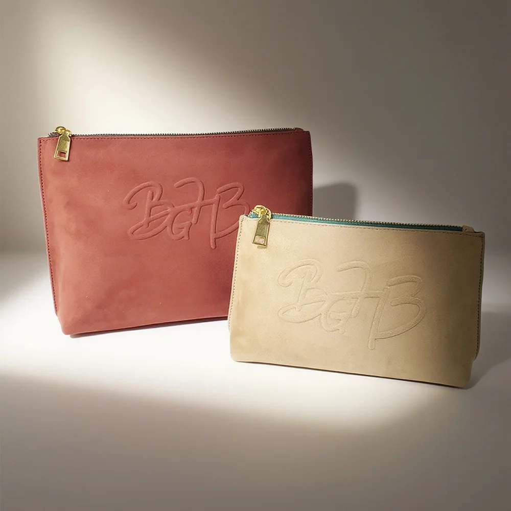 Two suede zip pouches, terracotta and champagne, with gold zipper pulls and debossed BGP logo, on lit neutral studio backdrop