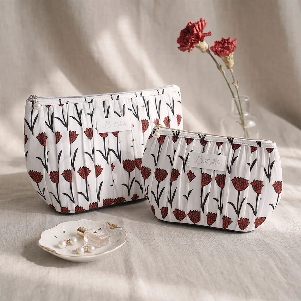 Two white pleated cotton pouches with red floral print and pearl zipper charms on beige linen, red carnations in glass vase behind, pearl earrings and perfume bottle on small ceramic dish in front